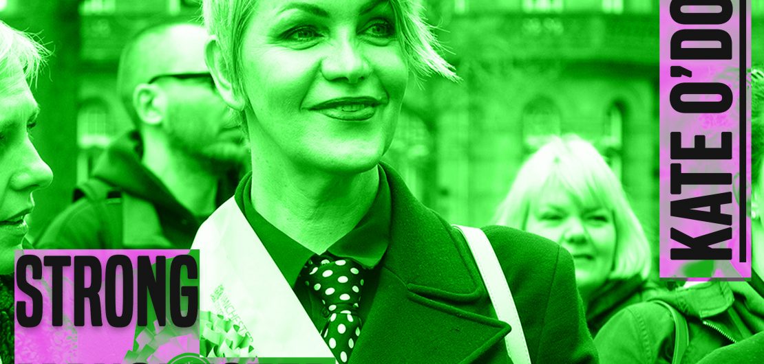 A woman stands in the front of a group of people. She has short, light hair, she is looking ahead and smiling. She is wearing a sash. The photo is from chest up.The image has a green hue to it. to the left of her knees are the words 'strong manchester women' in grey lettering and a thick font in capital letters. In the same style font, next to her right ear are the words 'Kate O'Donnell'