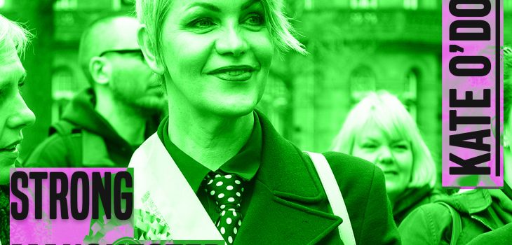 A woman stands in the front of a group of people. She has short, light hair, she is looking ahead and smiling. She is wearing a sash. The photo is from chest up.The image has a green hue to it. to the left of her knees are the words 'strong manchester women' in grey lettering and a thick font in capital letters. In the same style font, next to her right ear are the words 'Kate O'Donnell'