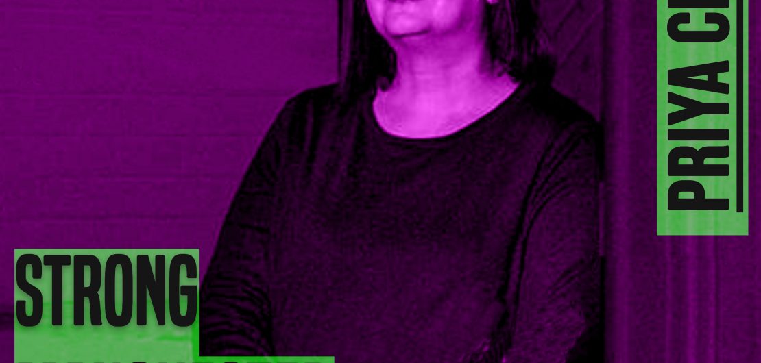A woman with dark hair sits on a chair and she is wearing a dark top. The photo crops above her knees. The picture has a purple hue to it. On the leff of the picture are the words 'strong manchester women' on the right 'Priya Chopra - both in capital and grey colour letters