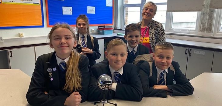4 kids in school uniform stand smiling in front of a microphone. A woman stands behind them and smiles