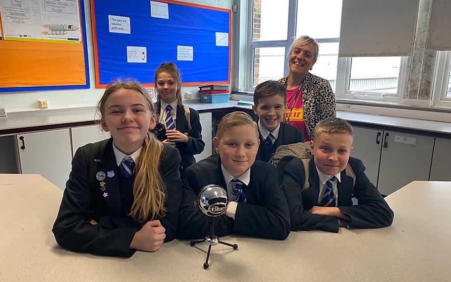 4 kids in school uniform stand smiling in front of a microphone. A woman stands behind them and smiles