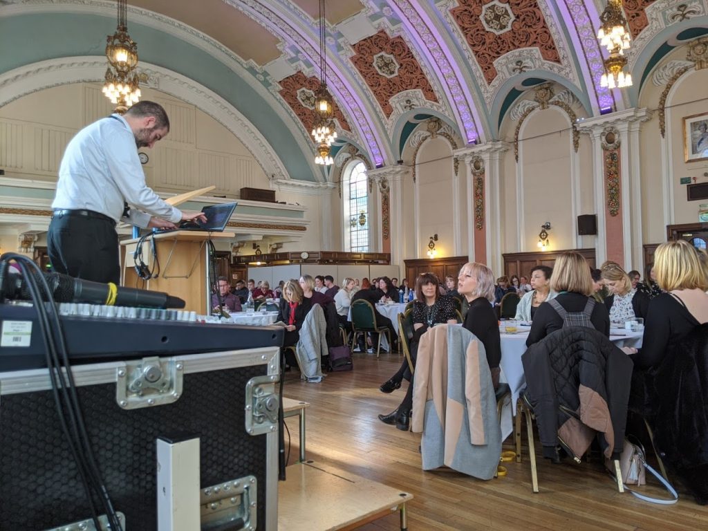 a photo of a room full of people sat at tables. The ceiling of the room is ornate and a mam stands at a lectern at the front of the room.