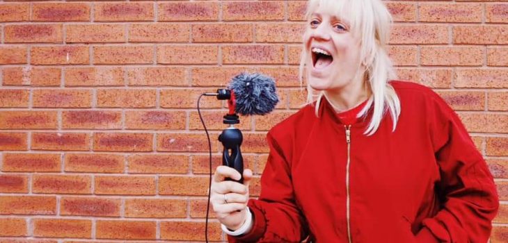 a women is standing in fron tof a brick wall, she is holding a microphone and speaking into it. she has a red coat on a and medium length blonde hair