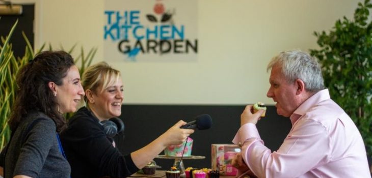 3 people sit around a table. One is talking, the other eating a cupcake and the other holding microphone