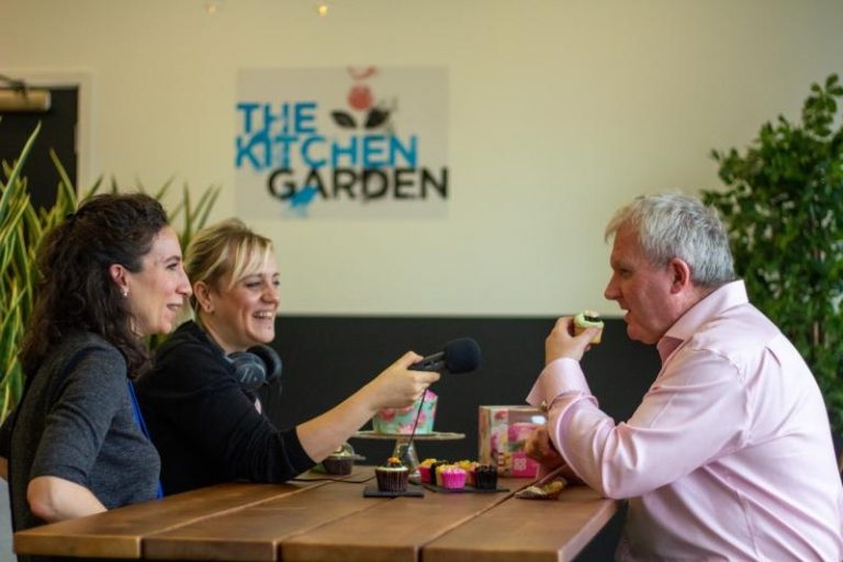 3 people sit around a table. One is talking, the other eating a cupcake and the other holding microphone