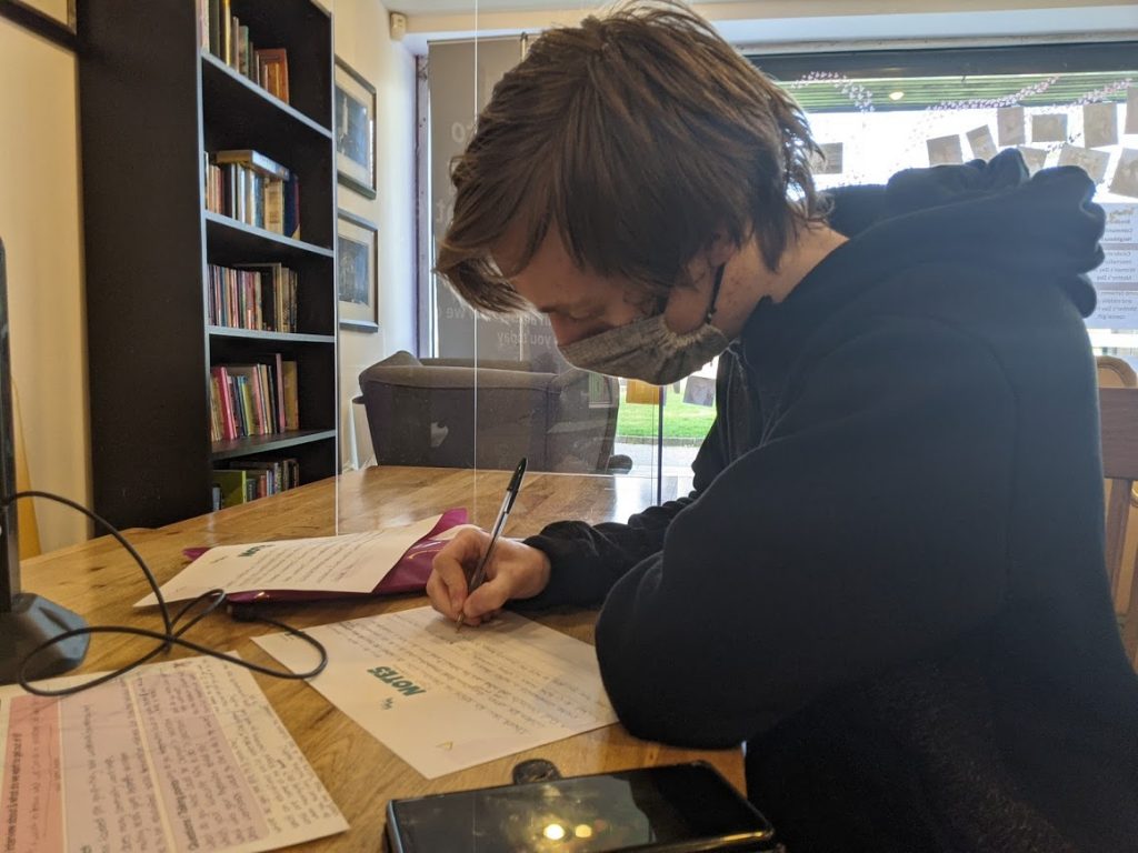 Image of young man in black jumper with a grey face mask on filling in script notes. 