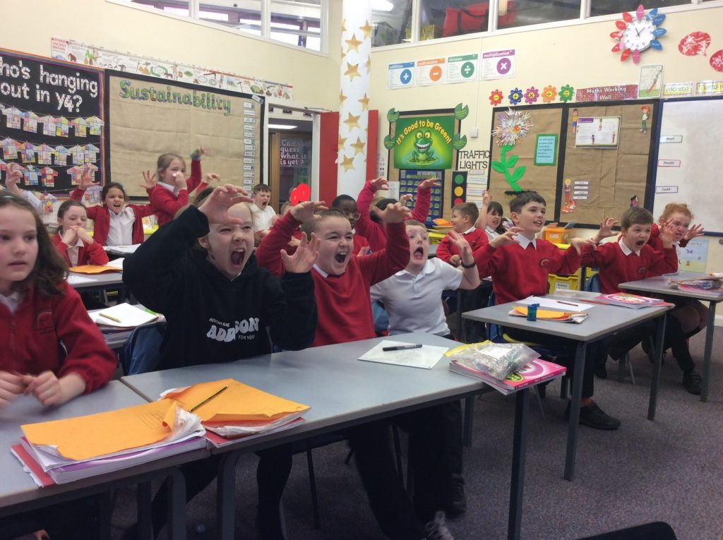 Image of primary school students in a classroom with their hands in the air